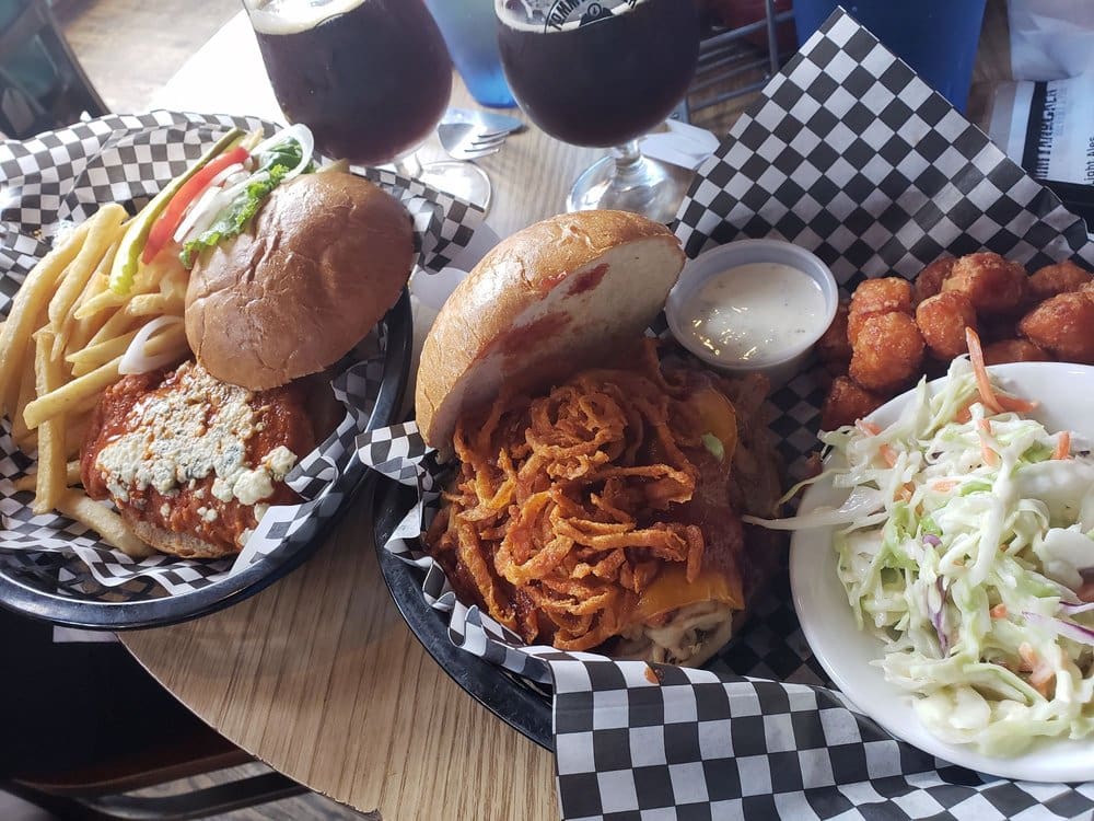 Two baskets of food, one with buffalo chicken patty and blue cheese with side of fries, the other with a burger with cheese, bacon, fried onions, and side of tater tots and cole slaw and side of two beers